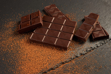 Close-up of broken chocolate pieces and cocoa powder on dark stone background with copy space.