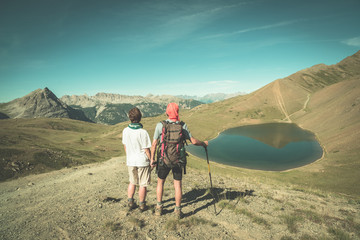 Fototapeta na wymiar Couple of hiker on the mountain top looking at blue lake and mountain peaks. Summer adventures on the Alps. Wide angle view from above, toned image, vintage style.