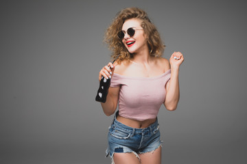 Cute stylish curly young woman drinking soda from the bottle over gray background