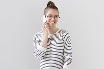 Indoor photo of nice young European female standing isolated against gray background in big round glasses, looking sideways and talking on cellphone with joyful expression on face and cheerful smile.