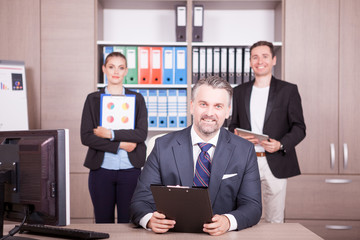 Businessman at the desk looking at a folder and his colleagues in background smiling