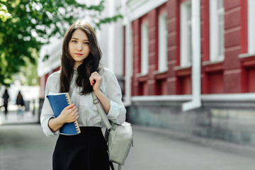 Beautiful girl student with a backpack and a textbook standing outdoors with the building of the University on the background
