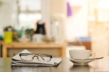 Closeup cup of hot cappuccino coffee with newspaper and spectacles on the wood table with blur kitchen room background-warm tone.