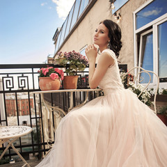 Young pretty stylish bride in fluffy white dress sitting at summer terrace. Beautiful young woman...