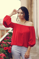 Young beautiful caucasian brunette woman with hairstyle posing at summer terrace with with on city buildings. Attractive girl in trousers and red blouse.