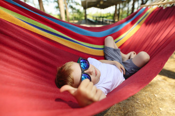 Fototapeta na wymiar Cheerful little boy with sunglasses lying on hammock in a pine forest, he make thumbs up gesture