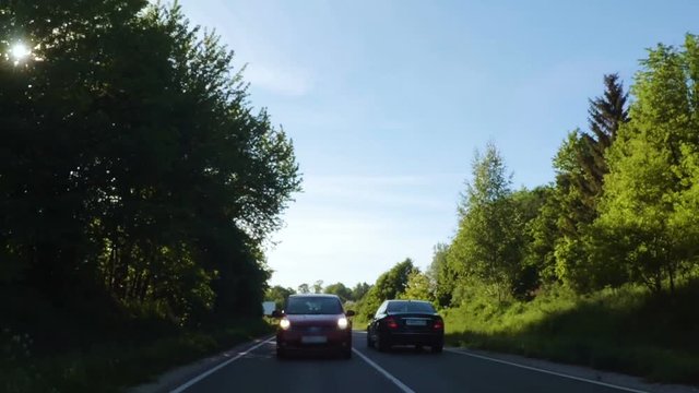 Travel around Europe by car. Mounted camera, back view. Camera view of driving on a long straight tarmac road towards sun in Europe. Trees on roadside of a two-lane road. Glare of  sun when driving.
