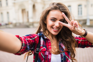 Young girl take selfie from hands with phone with victory sign on summer city street urban life...