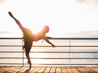 Beautiful ballerina performing pirouette. Blonde woman in black ballet tutu on embankment above ocean or sea beach at sunrise. Female dancing with arms. © kohanova1991