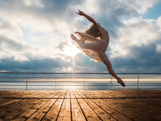 Jumping ballerina in beige dress and pointe on embankment above ocean or sea beach at sunrise.Beautiful blonde woman with long hair practicing stretching and classic exercises.Epic jump.