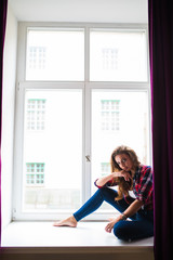 Fototapeta na wymiar Beautiful young woman looking through window while sitting at windowsill at home