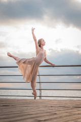 Obraz premium Dancing ballerina in beige silk dress and pointe on embankment above ocean or sea beach at sunrise. Young beautiful blonde woman with long hair practicing classic exercises with emotions.