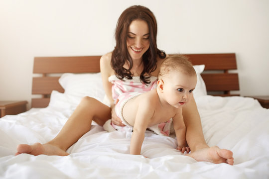 Young beautiful mom in sleepwear sitting on bed with her baby daughter smiling playing at home.