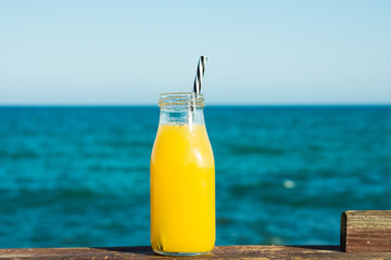 Glass bottle with citrus fruit juice orange tangerine with straw on wooden rail, turquoise sea and blue sky in the background, natural setting, vacation, summer, horizontal