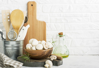 Fresh champignons in a wooden bowl on the background of a kitchen white brick wall with cutting boards, kitchen utensils, tools, olive oil for cooking dinner. Selective focus.