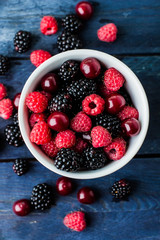 Top view of white cup with Summer fresh berries on blue wooden table background. Copy space. Vegetarian food - raspberry, blackberry and cherry. Rustic style