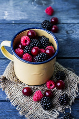 Summer fresh berries in yellow cup on blue wooden table background. Copy space. Vegetarian food - raspberry, blackberry and cherry. Rustic style