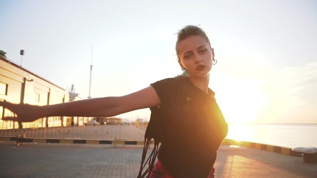 Young stylish funky girl with green hair riding roller skates and dancing near sea port during sunset, slow motion