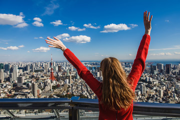 Happy young woman with her hands up. Girl stands with her back. Tokyo Tower with skyline on...