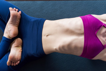 Close-up image of slim female body in sports bra and leggings lying on mat barefoot doing backbend...