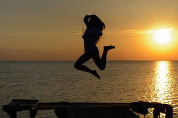 A girl in lingerie and shirt listening music on headphones on a pier by the sea at sunset