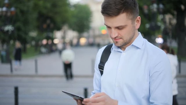 Handsome young man using tablet computer public space blurred city laptop ebook e-reader outside evening smiling tapping touching screen checking hands holding netbook blue shirt brunette urban blur