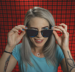 Portrait of a trendy hipster girl wearing glasses, winking at the camera, standing on a red checkered background. The concept of urban fashion. Turquoise shades