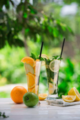 Detox water with fruits - orange and green apple and lemon and basil leaves and mint leaves on white table outdoors in summer garden. Homemade mojito or lemoade with ice cubes.