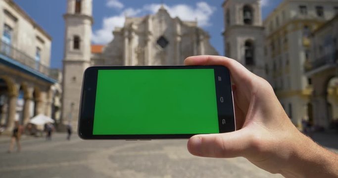 Holding and tapping a green screen smartphone in portrait mode outside Havana Cathedral. With optional luma matte.	 	