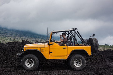 Fototapeta na wymiar A curly-haired man drinking water is sitting in the offroad yelow vehicle parked at the top of a valley with volcanic rock and mountains in Bali, Indonesia