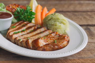 Barbecue pork steak slices on white plate. Delicious pork steak for lunch or dinner on wood table. Moist and soft homemade pork barbecue served with mash potato barbecue sauce and vegetable.