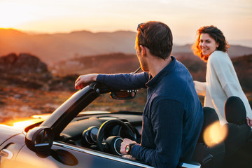 A young couple have fun in a convertible at sunset, around them beautiful nature - obrazy, fototapety, plakaty
