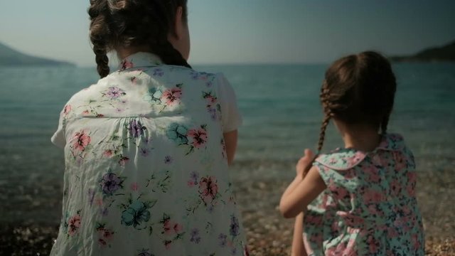 Two girls sit on the shore and throw stones into the water. Two pigtails are braided on the head of the sisters. Dressed babes in dresses with floral print. In sunny weather spend interesting time