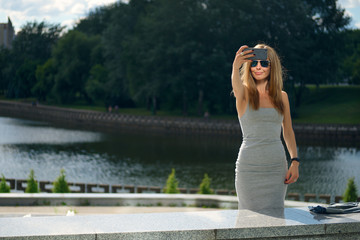 Attractive tourist girl making selfie with river bank on background in city park