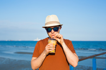 Portrait of cheerful attractive bearded hipster young man drinking iced coffee or frappe on the background of the sea
