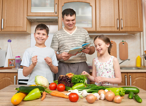 Father and two children reading cooking book and choice dishes. Happy family, girl and boy having fun with fruits and vegetables in home kitchen interior. Healthy food concept.