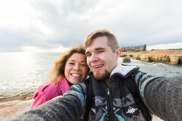 Young laughing couple hiking taking selfie with smart phone. Happy young man and woman taking self portrait with sea or ocean scenery on background. Winter time