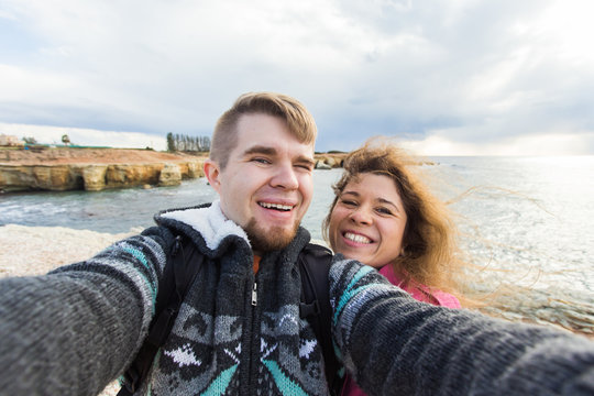 Young laughing couple hiking taking selfie with smart phone. Happy young man and woman taking self portrait with sea or ocean scenery on background. Winter time
