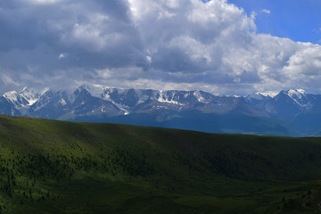 Fototapeta premium View of white peaks of North-Chuyski ridge and hills with green fields in Altai mountains in cloudy weather. Aktash, Altay Republic, Russia.