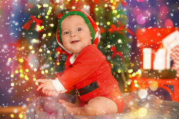 Cute baby in costume of Santa's helper and Christmas tree on background. Holidays celebration concept