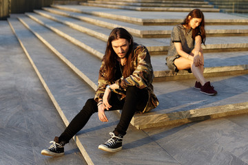 People, love and relationships problems concept. Outdoor shot of sad young couple not talking to each other after fight. Gloomy hipster sitting on concrete stairs ignoring his girlfriend in background