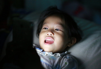 Happy Kid girl playing smartphone lying on a bed at night.
