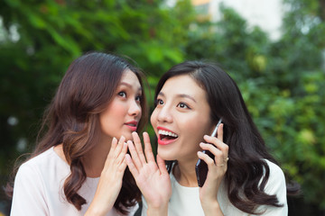Young woman whispering into cheerful friend's ear while on call