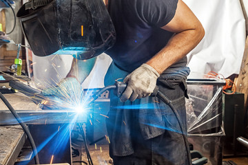 A man welder in a black T-shirt, construction gloves and a welding mask is enthusiastically working...