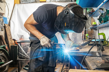 A man welder in a black T-shirt, construction gloves and a welding mask is enthusiastically working...