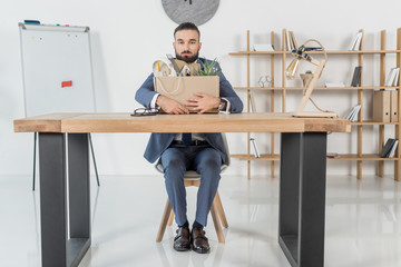 fired upset businessman sitting at workplace with cardboard box with office supplies