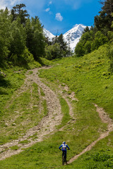 Fototapeta premium A lone biker climbs a mountain path up the mountain through an alpine meadow on a background of snowy mountains