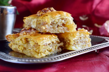Traditional Greek baklava made with filo dough, sugar syrup and wallnuts on a metal tray with metal coffee cup on a purple cloth background