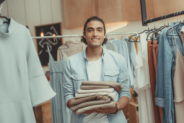 young business owner holding pile of pants while working in boutique before opening
