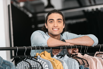 handsome young boutique owner leaning at hangers with clothes and smiling at camera